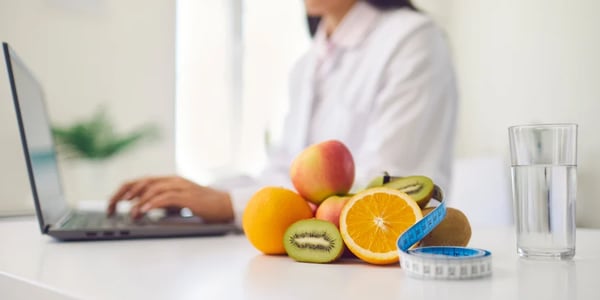 Mulher nutricionista vestida de jaleco branco, utiliza notebook sentada em uma mesa, com uma pilha de frutas e uma fita métrica ao lado.