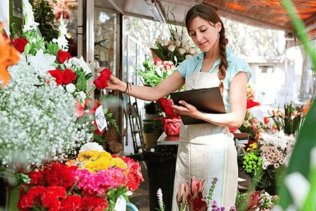 Uma floriculturista em pé de avental, com uma prancheta na mão, enquanto cuida das rosas.