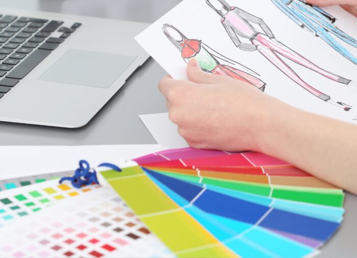 Na foto, uma mão segura um papel com croquis de roupas, ao lado um cartela de coloração pessoal e ao fundo o teclado de um notebook.