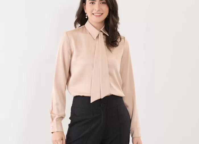 Modelo veste blusa com gravatinha veridiana dash, feita em cetim na cor areia.